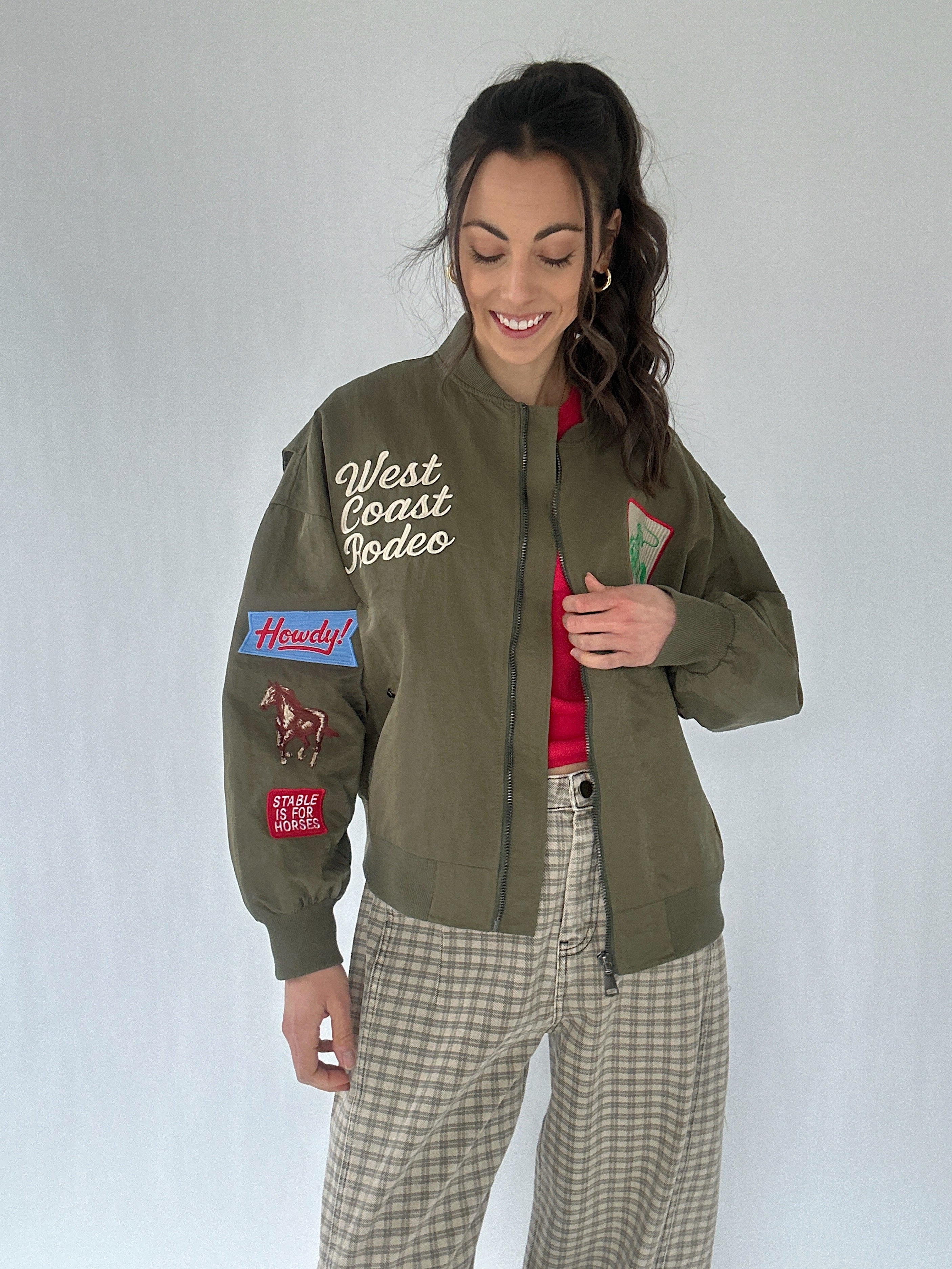 Woman wearing a green jacket with patches and text, standing against a plain background