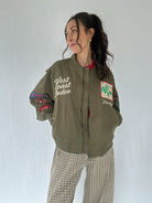Woman wearing a green bomber jacket with patches and text, standing against a plain background