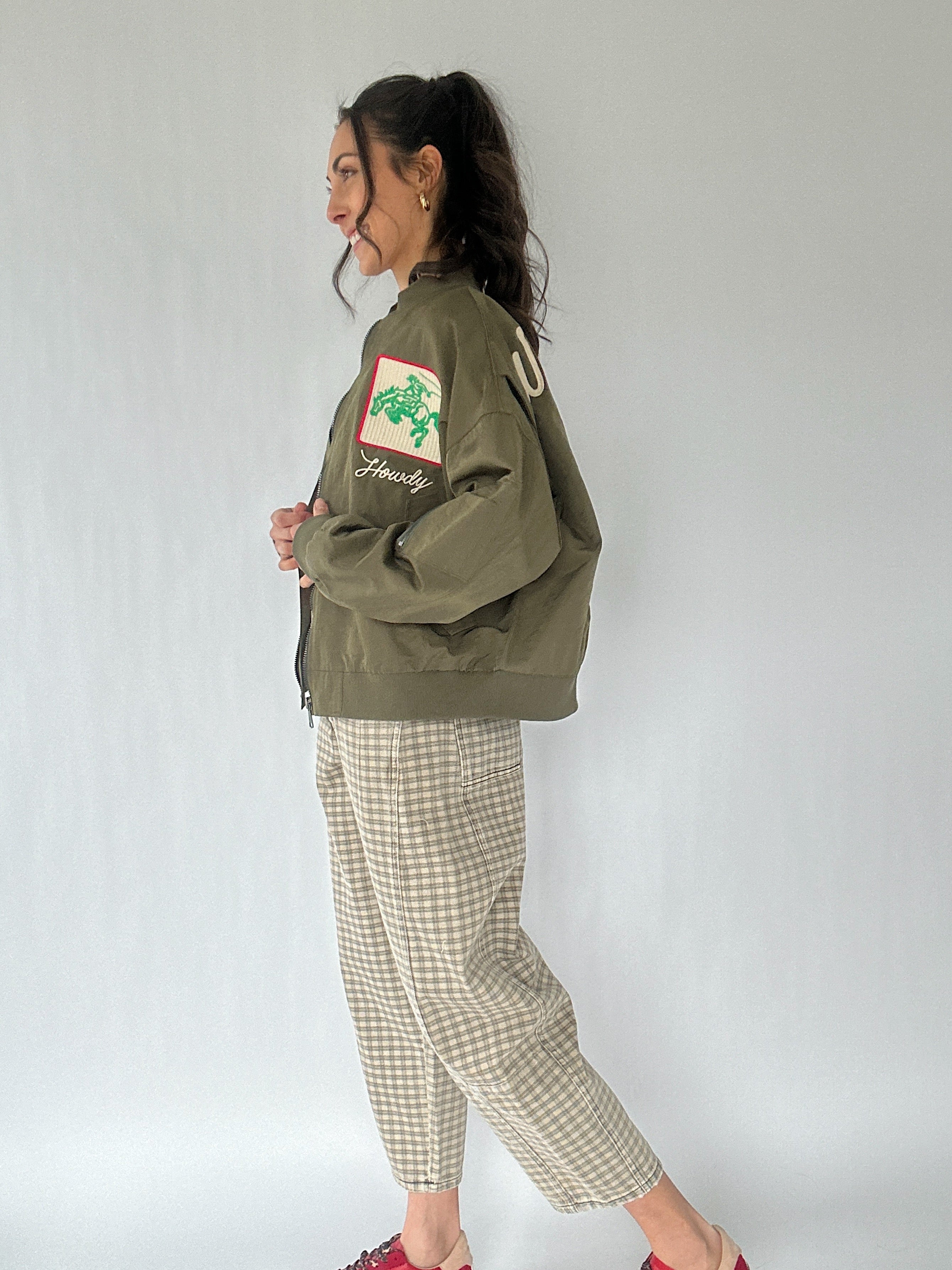 Woman wearing a green jacket with a logo and text, standing against a plain background from side