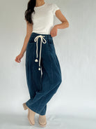 Person wearing blue corduroy barrel drawstring pants with a white top on a plain background with legs crossed