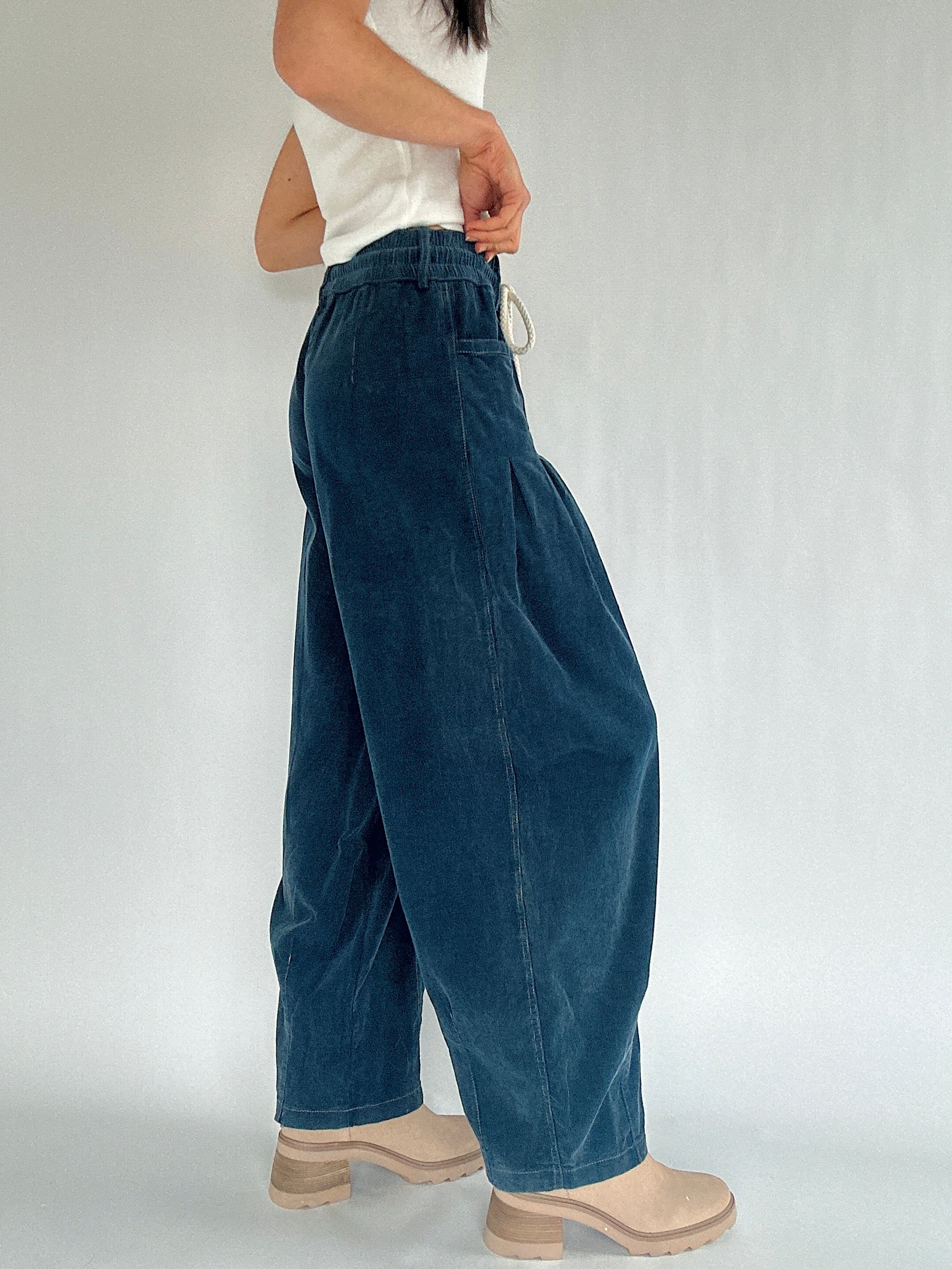 Person wearing blue corduroy barrel wide-leg pants and a white top on a plain background from the side