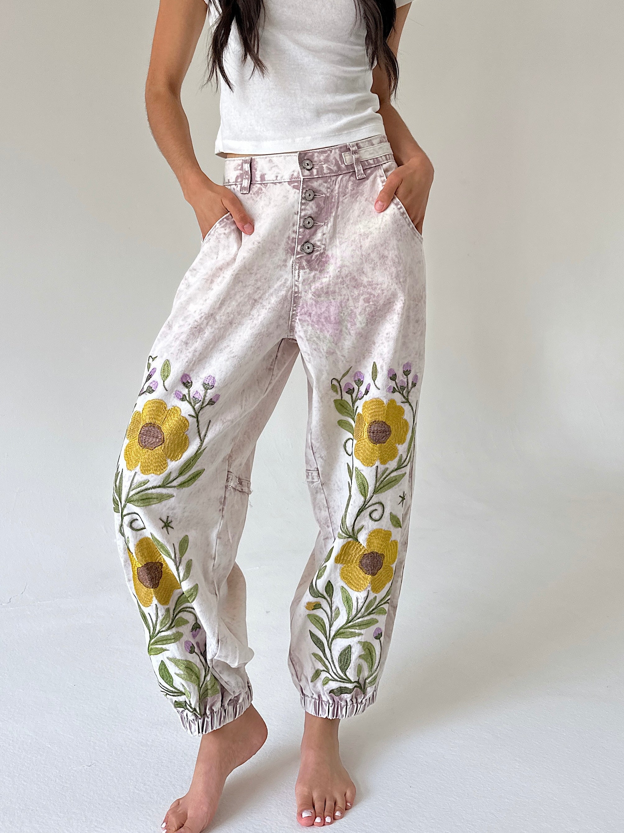 A person wearing loose, high-waisted jogger-style pants with a light beige and lavender tie-dye pattern, yellow floral embroidery on the lower legs, and elastic cuffs, standing barefoot against a plain light background.