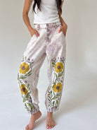 A person wearing loose, high-waisted jogger-style pants with a light beige and lavender tie-dye pattern, yellow floral embroidery on the lower legs, and elastic cuffs, standing barefoot against a plain light background.