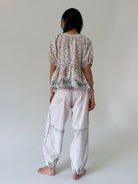 A woman shown from the back wearing a short-sleeve floral peplum top and loose light gray pants, standing barefoot against a plain light gray studio background.