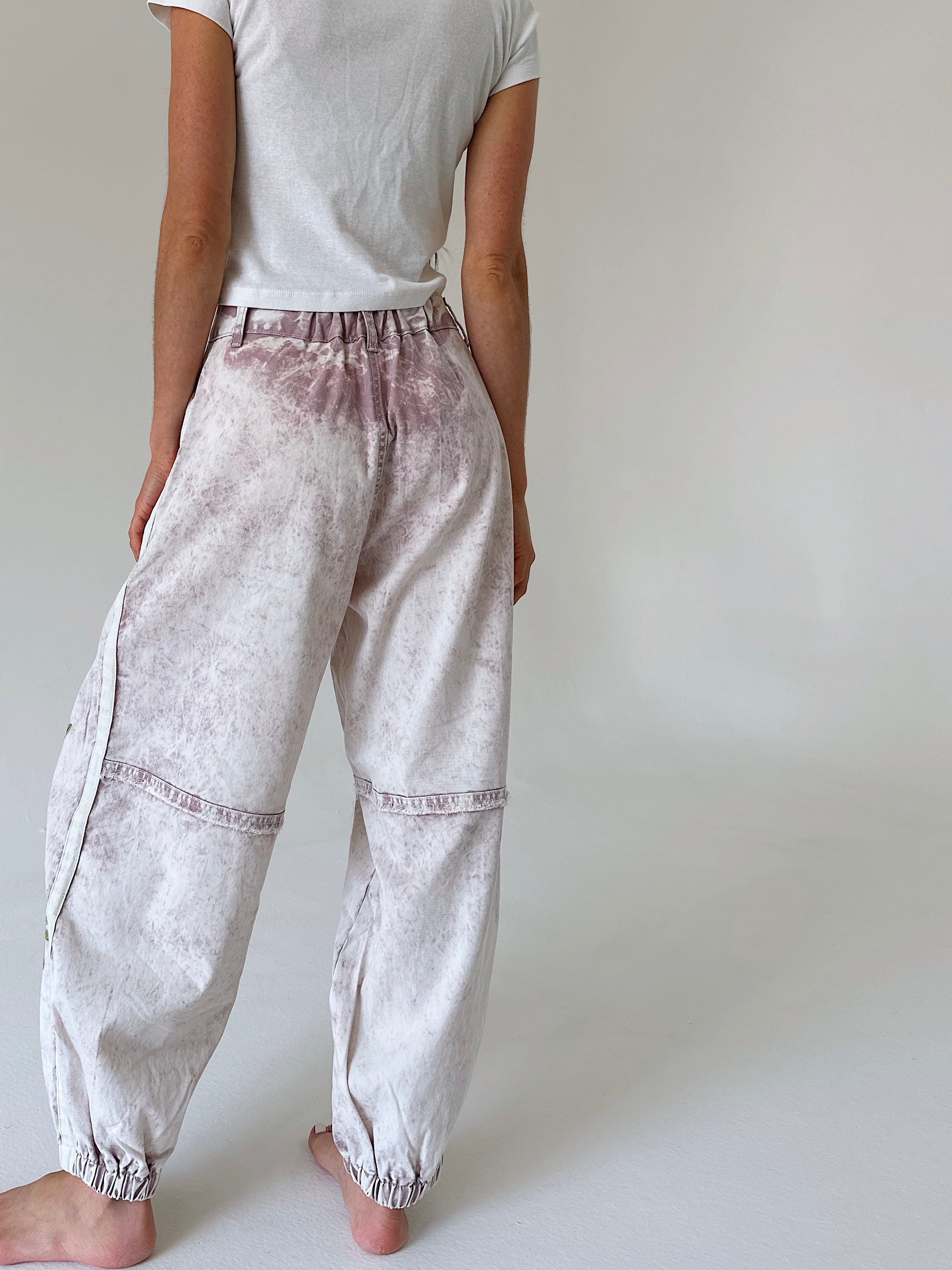 A person shown from the back wearing loose, elastic-cuffed pants with a faded dusty mauve and off-white tie-dye pattern, paired with a white t-shirt against a plain light gray studio background.