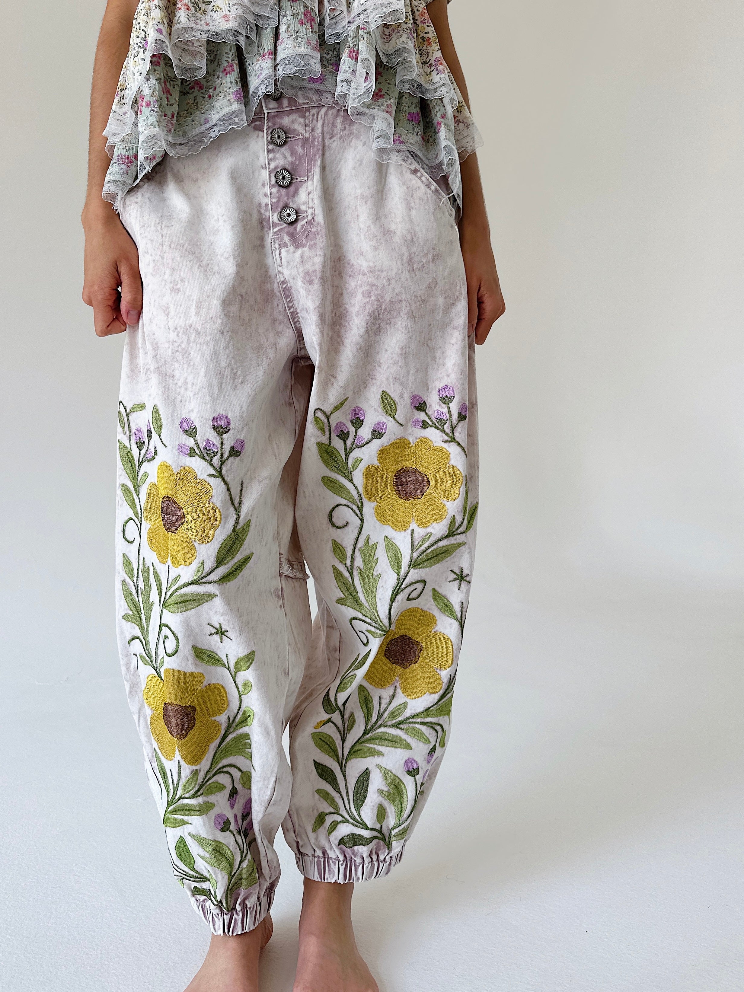 A person wearing loose, light pink-colored floral embroidered pants with yellow flowers and green leaves, paired with a ruffled top, standing against a plain light background.