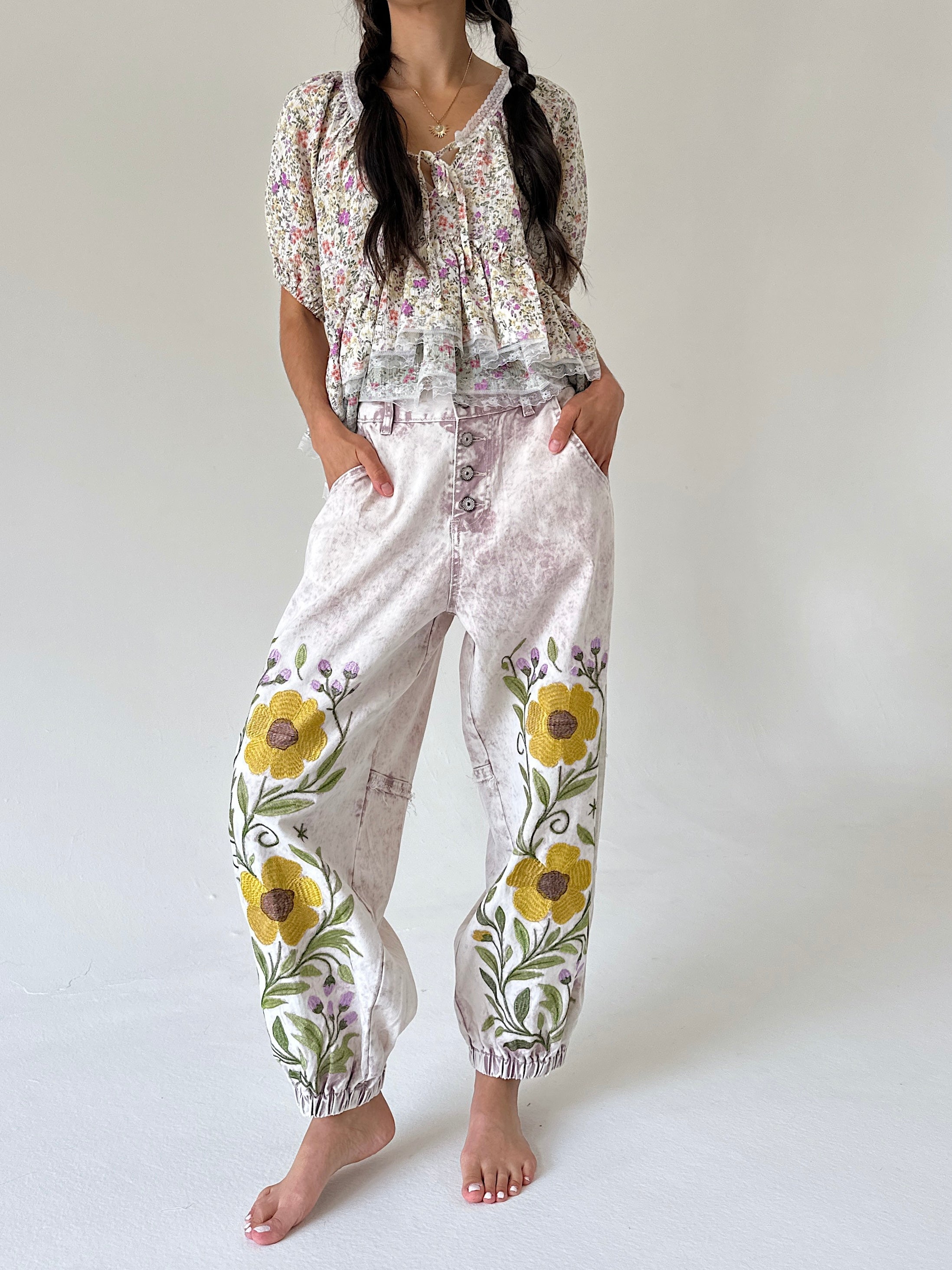 A woman modeling a bohemian-style two-piece outfit featuring a floral print top and matching pants with large yellow flower and green leaf embroidery on the legs, set against a plain light gray background.