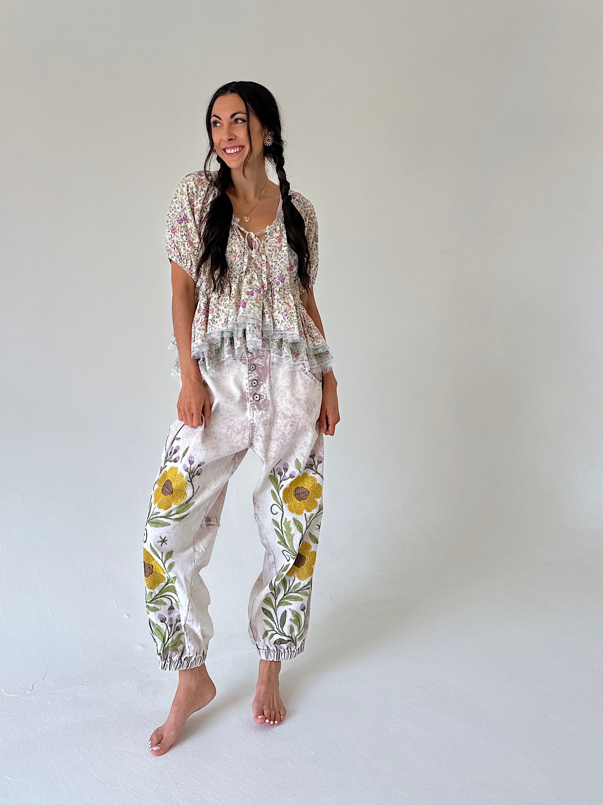 A woman modeling a boho outfit featuring a short-sleeve tie-front floral top and relaxed pants with yellow floral embroidery, set against a plain light gray studio background.