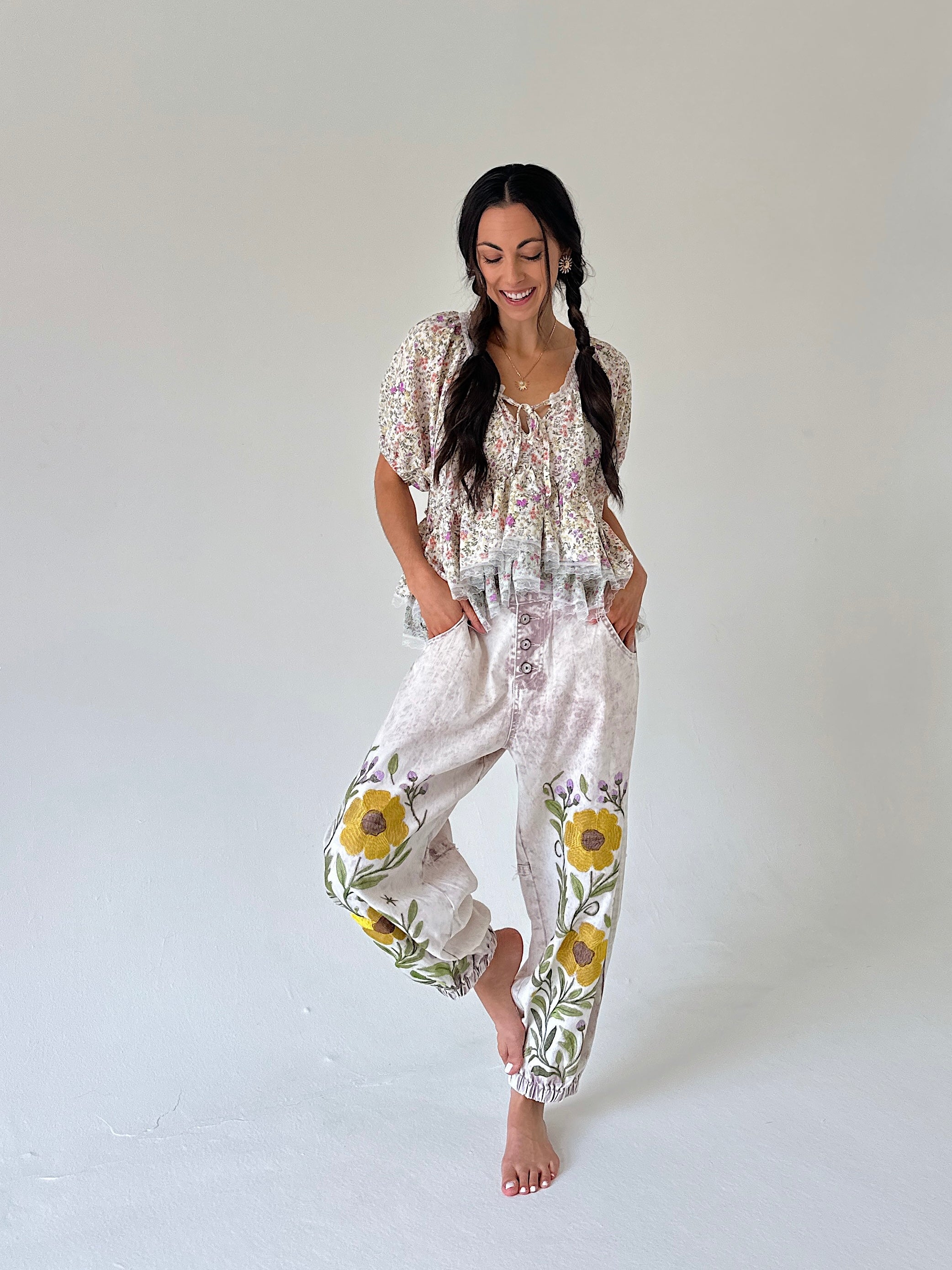 A woman modeling a bohemian-style two-piece outfit consisting of a short-sleeve tie-front top and matching loose pants with yellow floral embroidery, set against a plain light gray studio background.