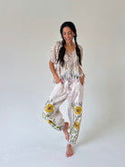 A woman modeling a bohemian-style two-piece outfit consisting of a short-sleeve tie-front top and matching loose pants with yellow floral embroidery, set against a plain light gray studio background.