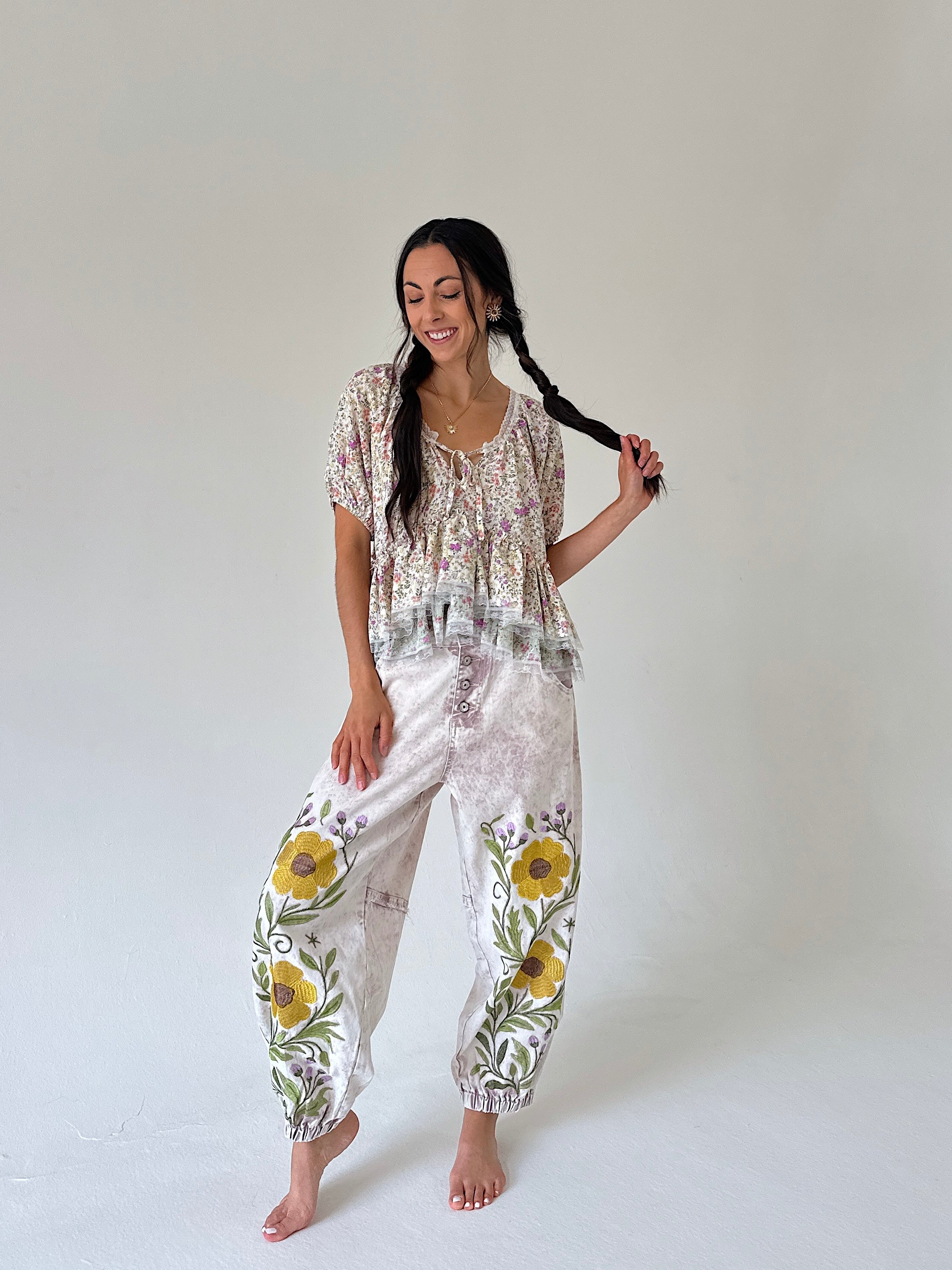 A woman modeling a bohemian-style two-piece outfit consisting of a short-sleeve floral top and printed pants with yellow sunflower embroidery, set against a plain light gray studio background.