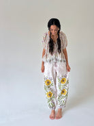 A woman stands barefoot against a plain light gray studio background, wearing a loose floral two-piece outfit with a short-sleeve top and matching pants featuring large yellow and purple flowers with green leaves.