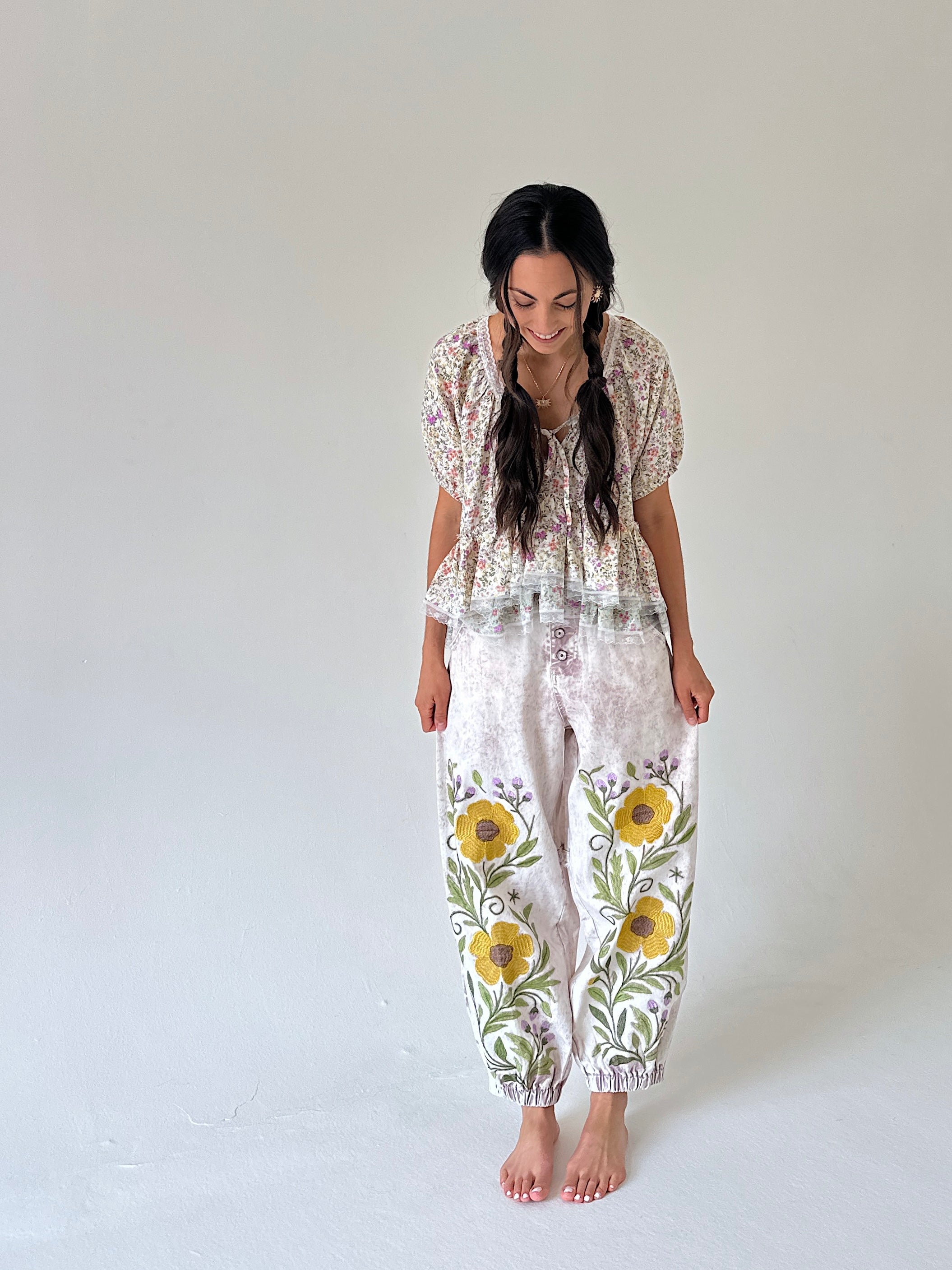 A woman stands barefoot against a plain light gray studio background, wearing a loose floral two-piece outfit with a short-sleeve top and matching pants featuring large yellow and purple flowers with green leaves.