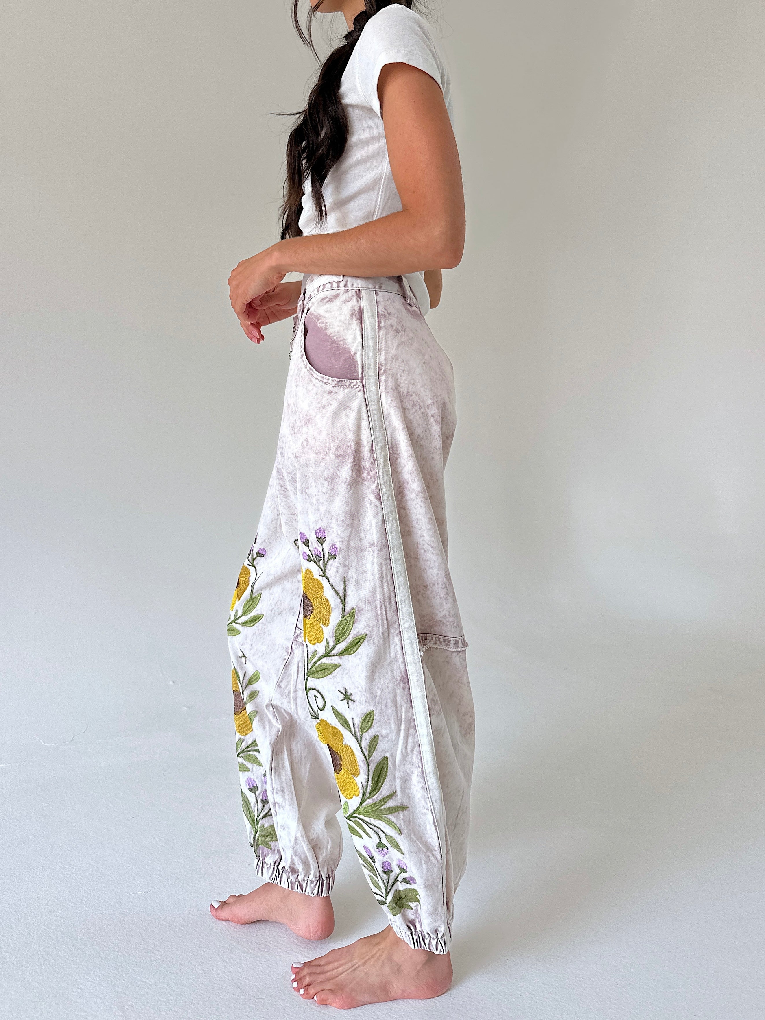 A woman wearing loose, high-waisted light pink pants with a yellow and green floral embroidery, paired with a white t-shirt, shown from a side profile against a neutral studio background.