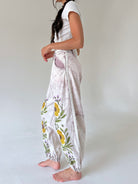 A woman wearing loose, high-waisted light pink pants with a yellow and green floral embroidery, paired with a white t-shirt, shown from a side profile against a neutral studio background.