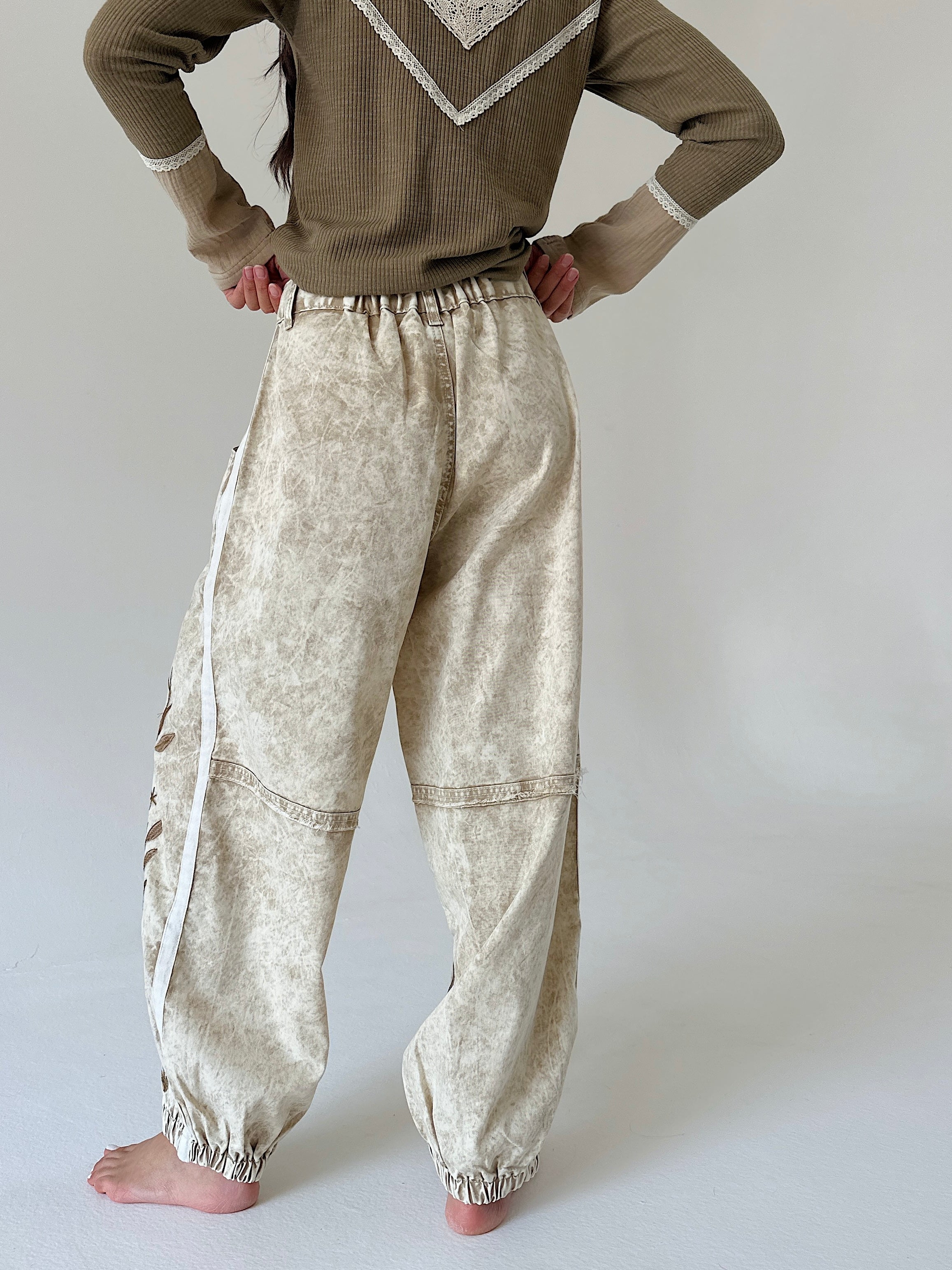 A person wearing loose, wide-leg beige pants with a subtle marbled texture and white side stripes, paired with a brown sweater, standing barefoot against a light gray studio background.