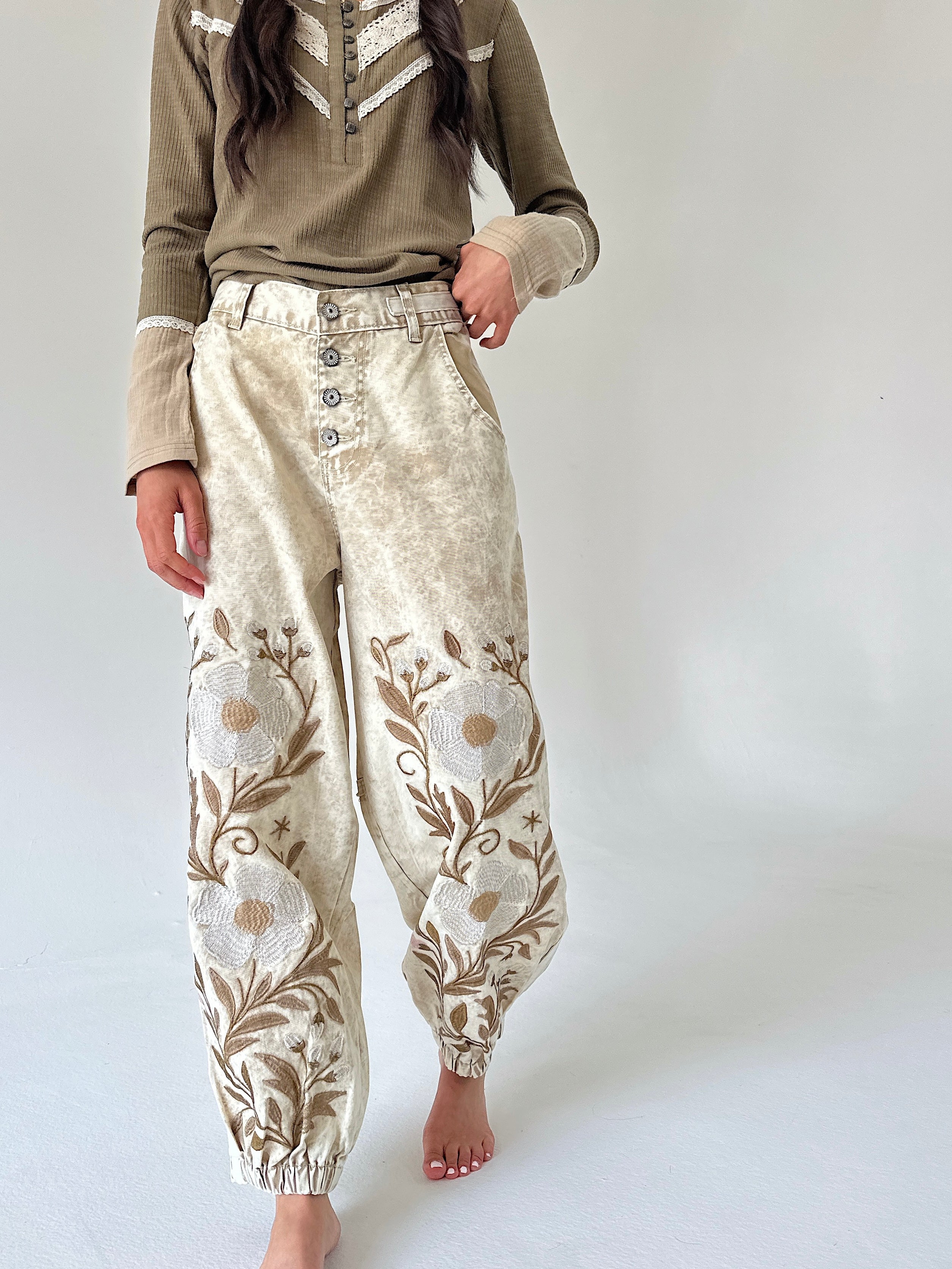 A person wearing loose, high-waisted cream and tan floral embroidered pants with a button fly, paired with an olive-brown long-sleeve top, standing barefoot against a plain light gray studio background.