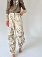 A person wearing loose, high-waisted cream and tan floral embroidered pants with a button fly, paired with an olive-brown long-sleeve top, standing barefoot against a plain light gray studio background.