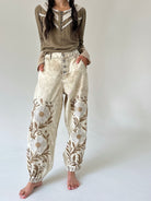 A model wearing a brown long-sleeve top with a lace-trimmed collar and cream-colored, high-waisted harem pants featuring a brown embroidered floral vine pattern, standing barefoot against a light gray studio background.