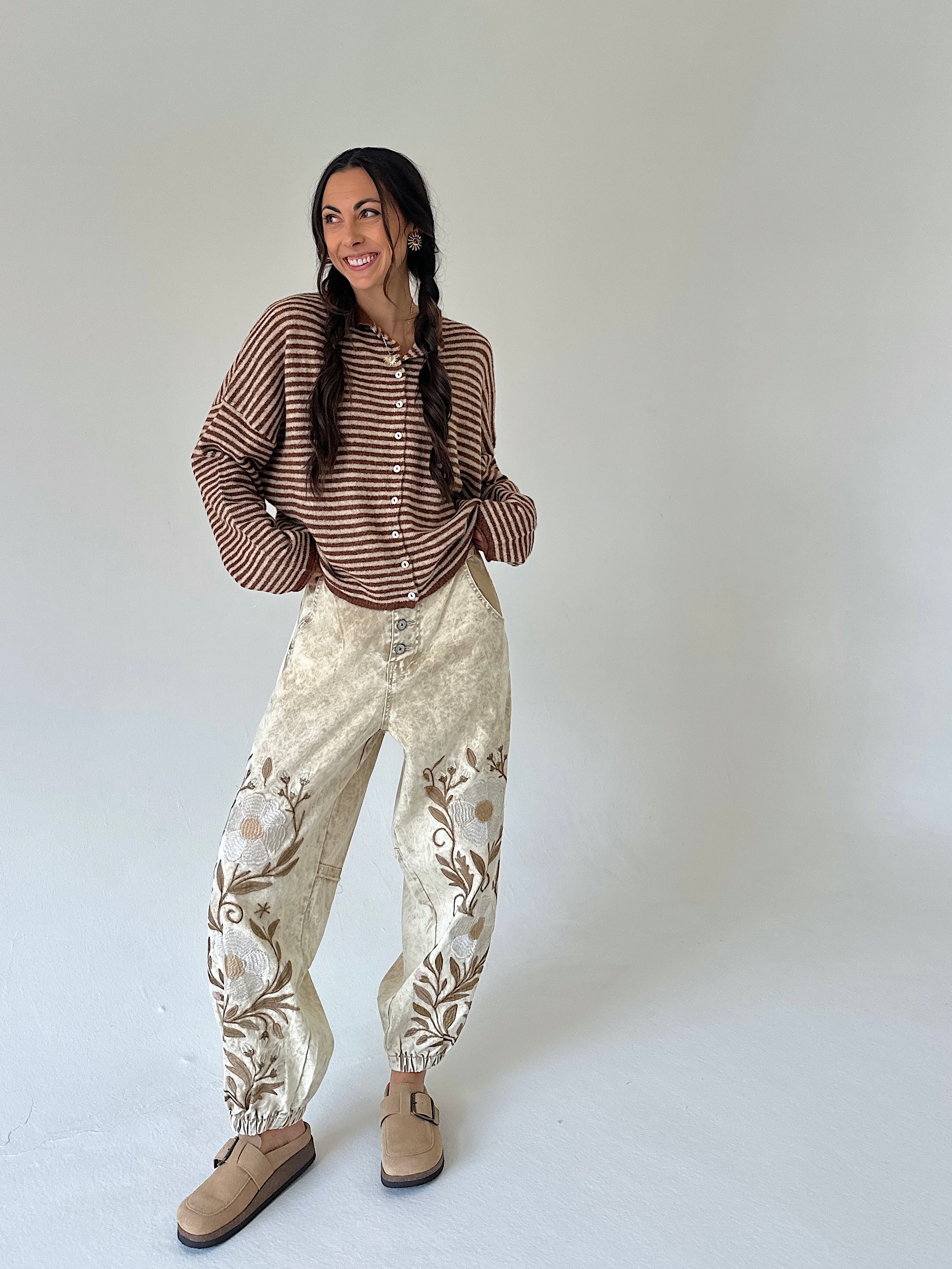 A woman modeling a brown and beige striped long-sleeve cardi paired with cream-colored pants featuring brown floral embroidery, styled with beige suede mules against a plain light gray studio background.