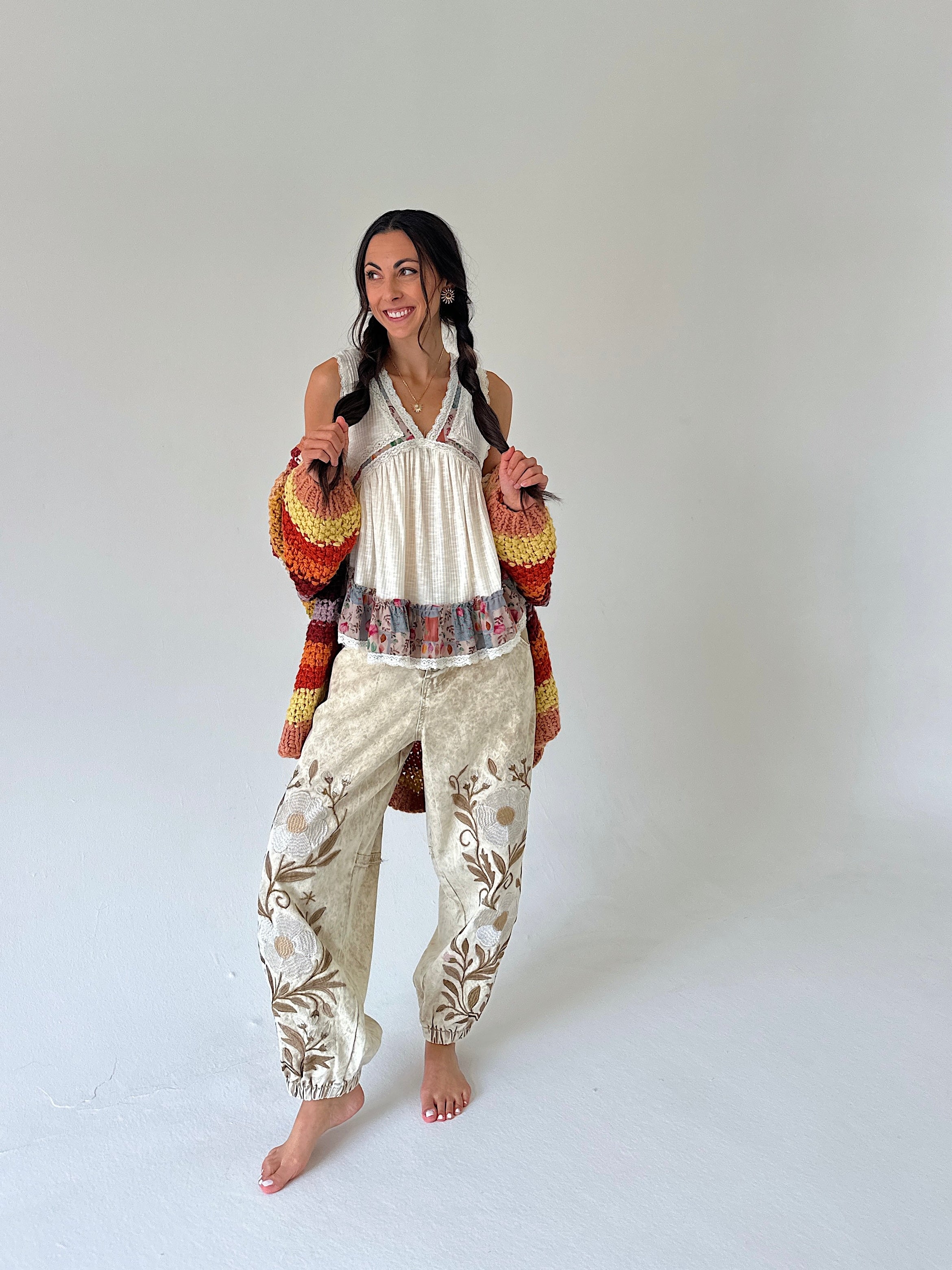 A woman modeling a bohemian-style outfit consisting of a cream pleated top, beige floral-print pants, and a multicolor knit cardigan, standing against a plain light gray studio background.
