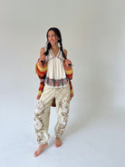 A woman modeling a bohemian-style outfit consisting of a cream pleated top, beige floral-print pants, and a multicolor knit cardigan, standing against a plain light gray studio background.