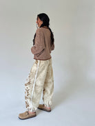 A woman stands in profile wearing a brown and beige striped long-sleeve top, beige patterned wide-leg pants with a white side stripe, and tan slip-on shoes against a plain light gray studio background.