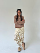 A woman stands against a plain light gray studio background wearing a brown and cream striped long-sleeve top, cream embroidered jogger style pants, and tan slip-on shoes.