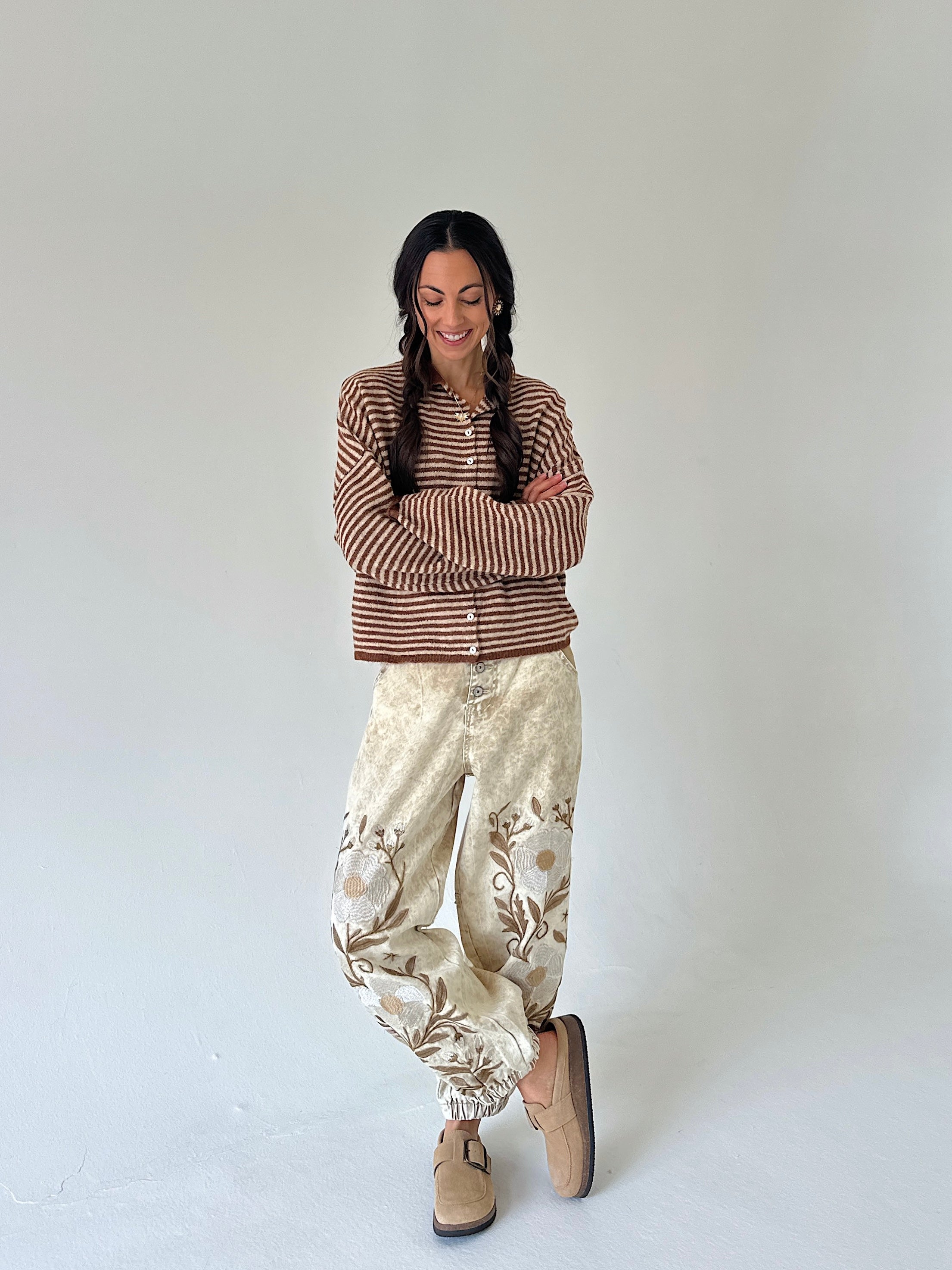A woman stands against a plain light gray studio background wearing a brown and cream striped long-sleeve top, cream embroidered jogger style pants, and tan slip-on shoes.