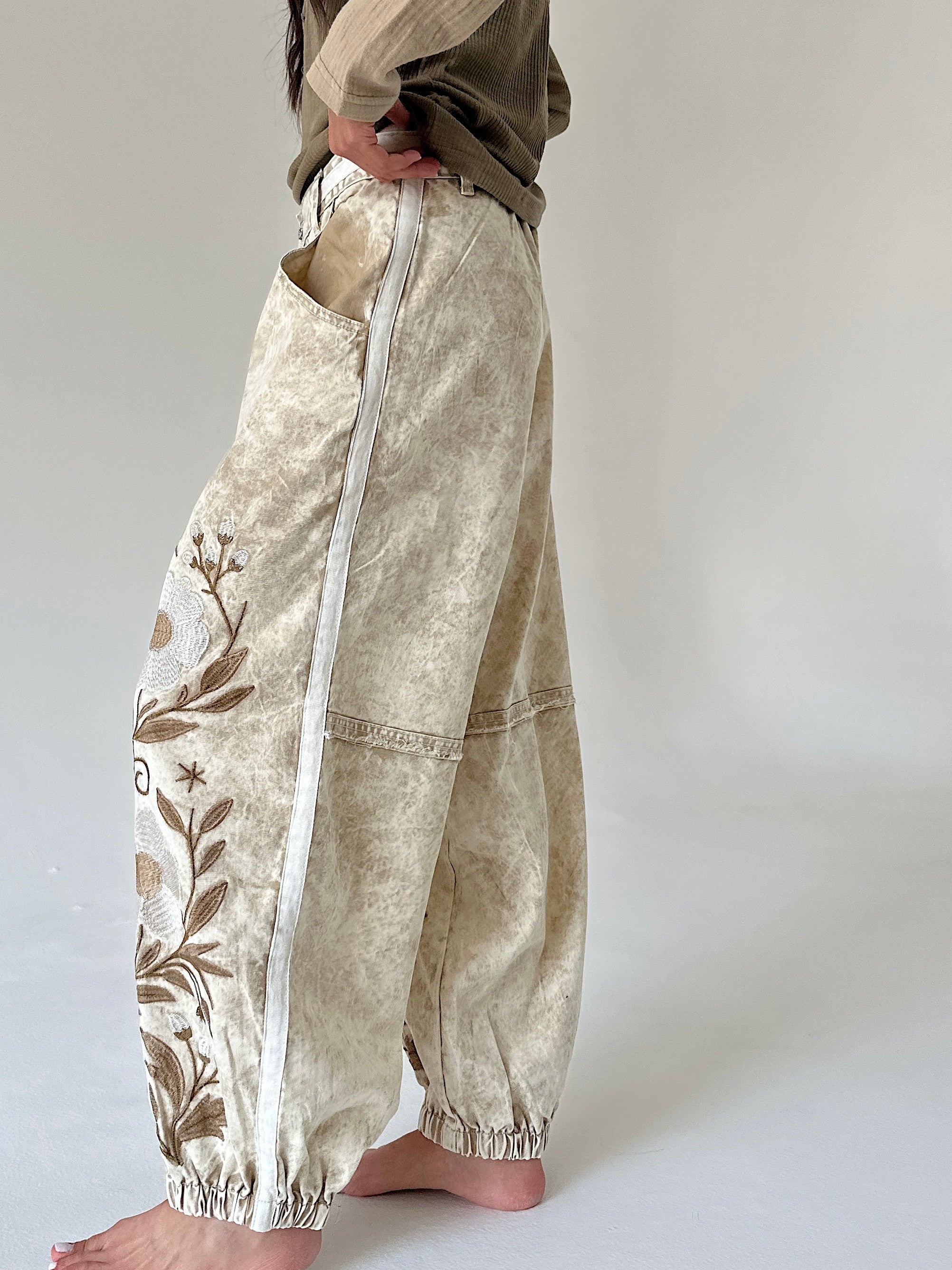 A person wearing loose, wide-leg cropped pants with a cream and beige marbled texture, brown floral embroidery on one leg, and a white vertical zipper detail, standing barefoot against a plain light gray studio background.