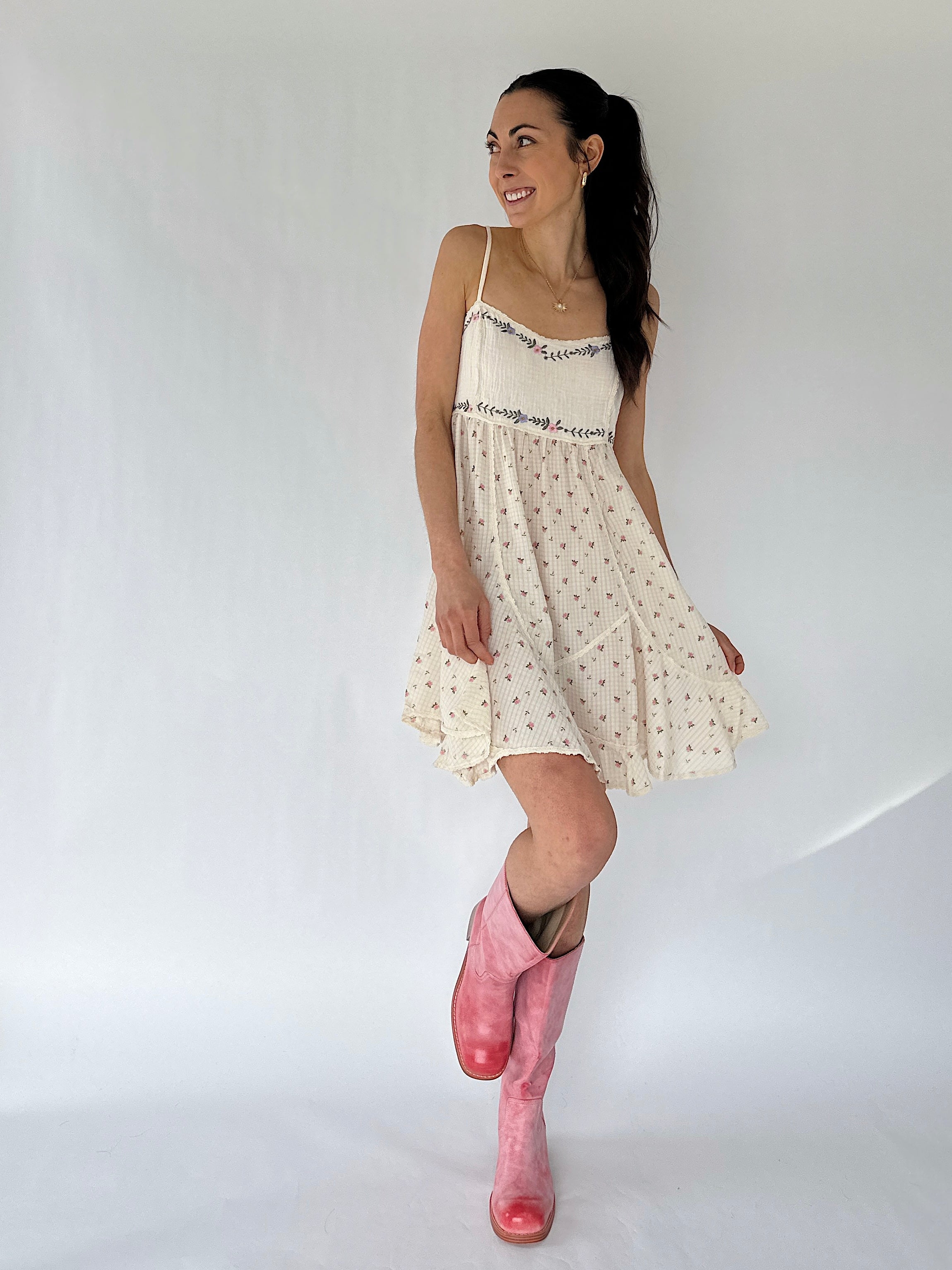 Woman wearing a white dress with floral patterns and pink boots on a plain background