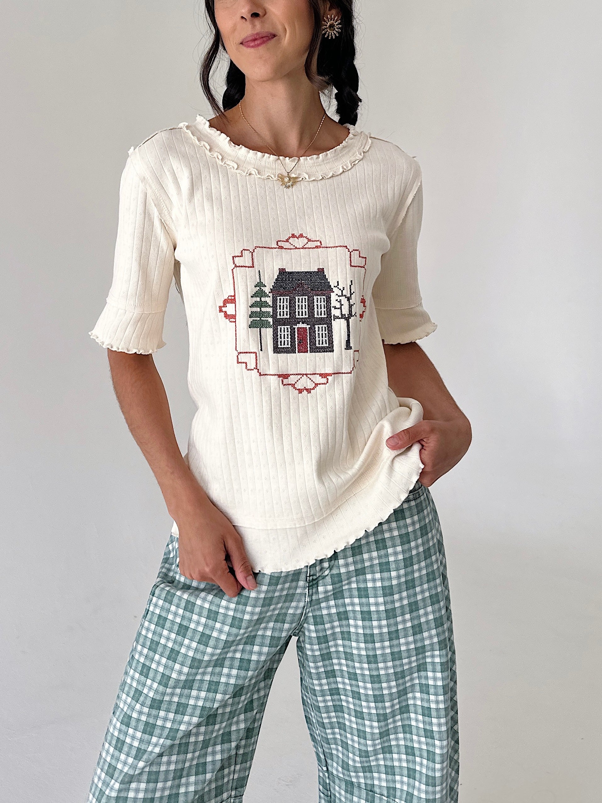 A woman wearing a cream ribbed short-sleeve top with a house embroidery graphic on the chest, paired with green and white plaid pants, against a plain light gray background.