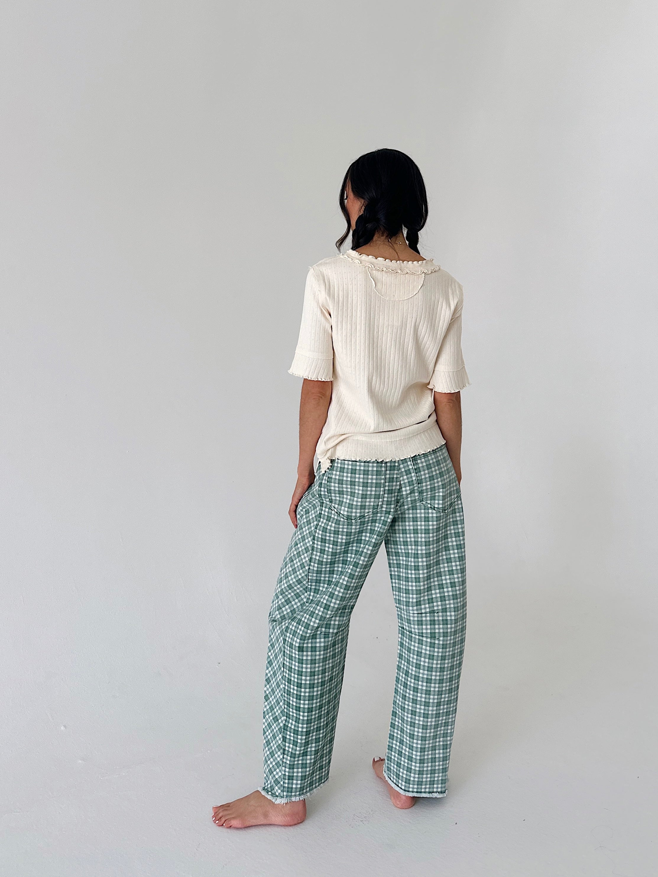 A woman stands barefoot against a plain light gray studio background, viewed from the back. She is wearing a cream ribbed short-sleeve top and green-and-white plaid pants.