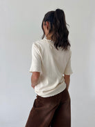A woman shown from the back wearing a cream ribbed short-sleeve top with ruffled trim and brown wide-leg pants, standing against a plain light gray wall.