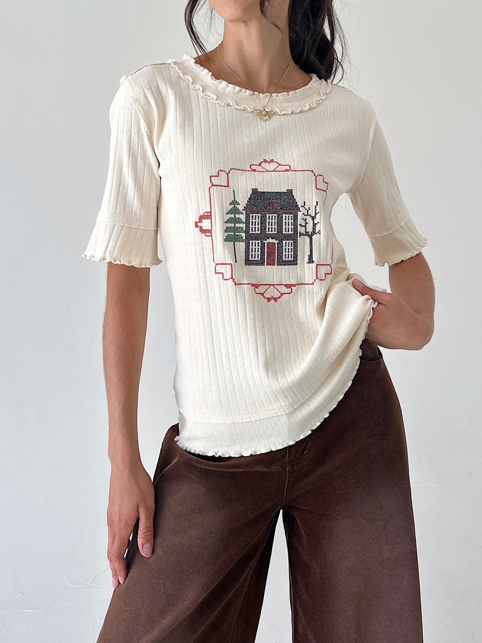 A woman wearing a cream ribbed short-sleeve top with a central house embroidery, paired with brown wide-leg pants, against a plain light gray background.