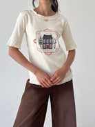 A woman wearing a cream ribbed short-sleeve top with a ruffled neckline and a central embroidered house graphic, paired with brown pants, against a plain light gray wall.