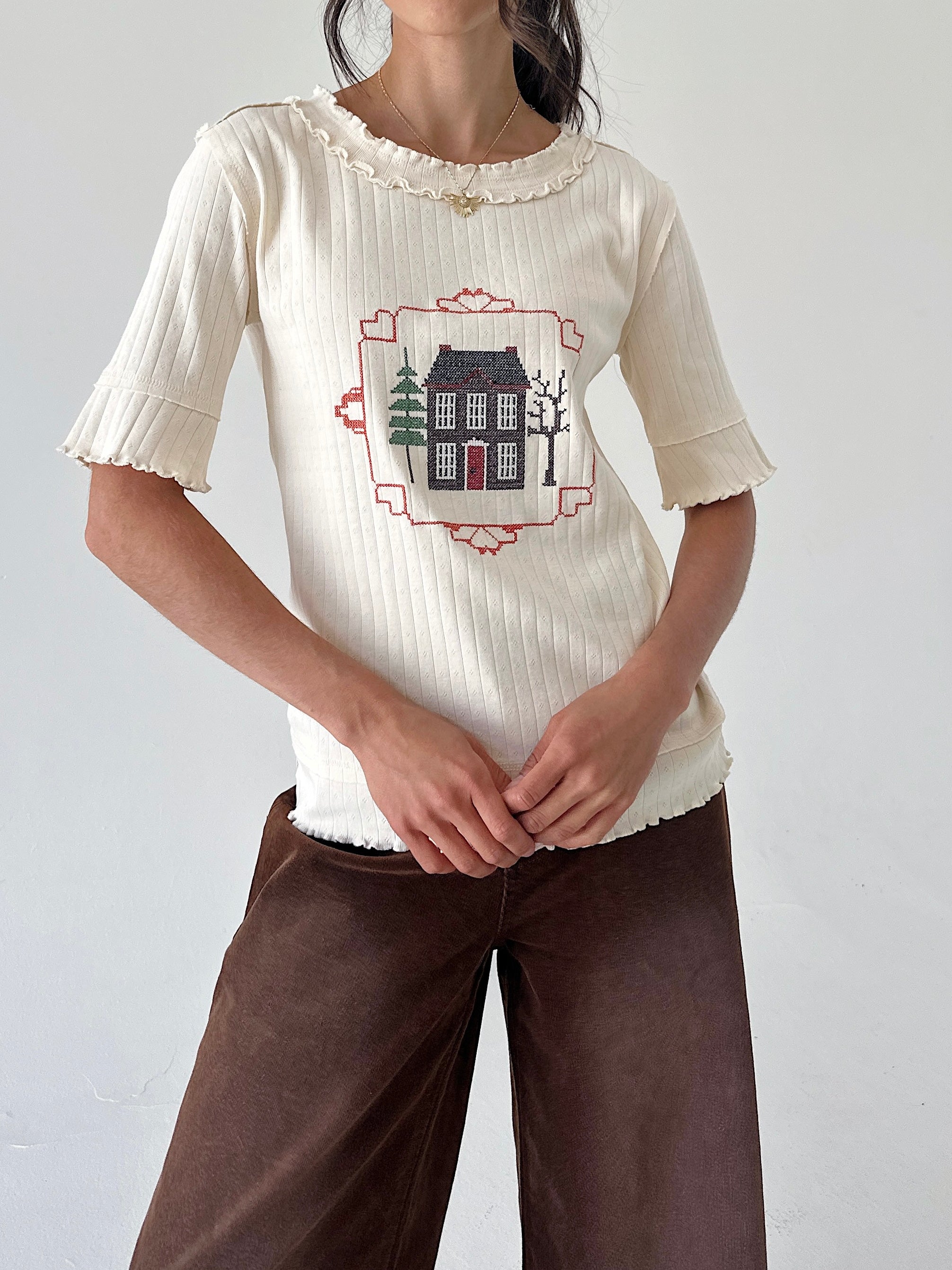 A woman wearing a cream ribbed short-sleeve top with a ruffled neckline and a central embroidered house graphic, paired with brown pants, against a plain light gray wall.