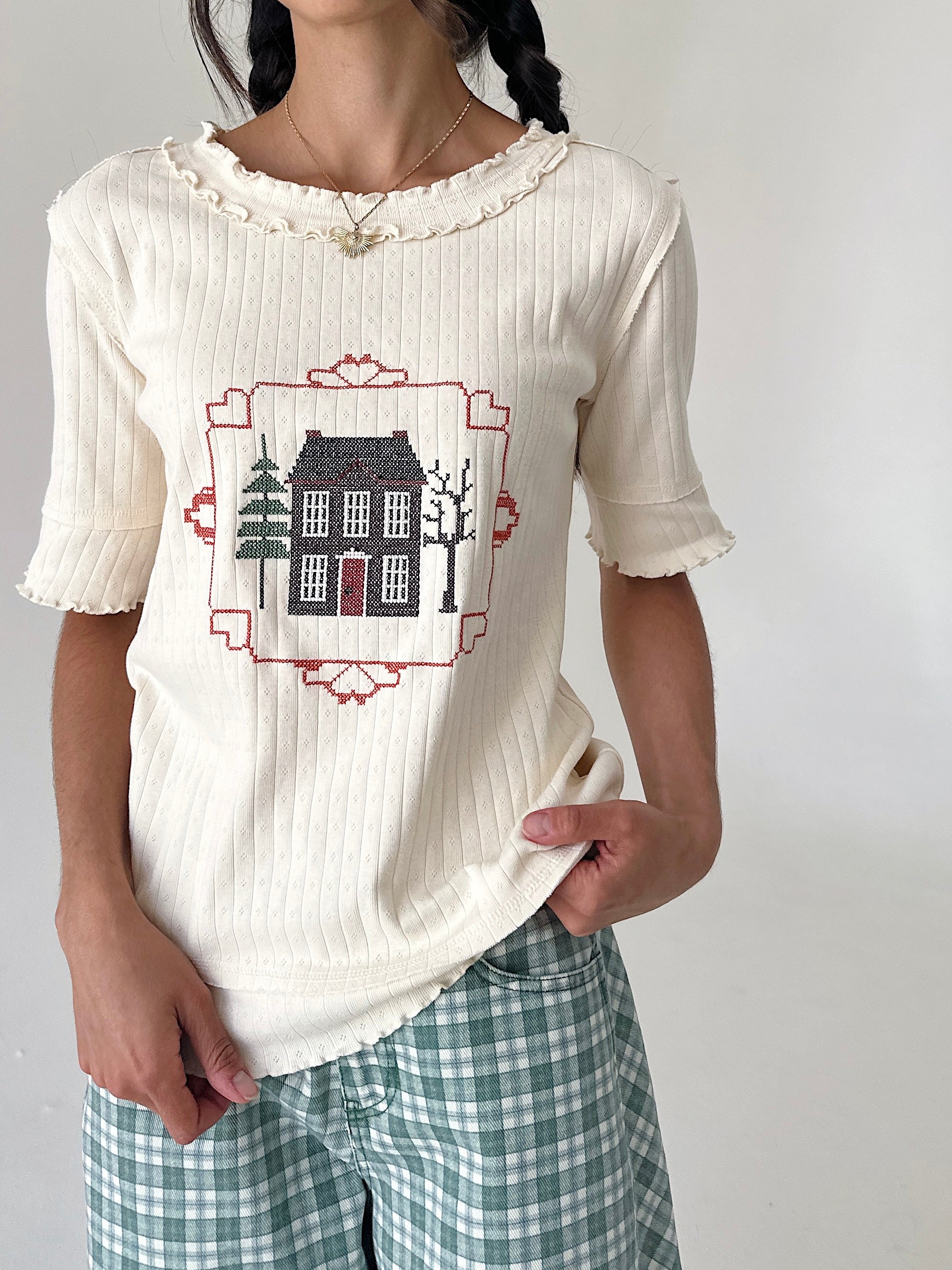 A woman wearing a cream ribbed short-sleeve top with a ruffled collar and a central embroidered house graphic, paired with green and white gingham pants, against a plain light gray background.