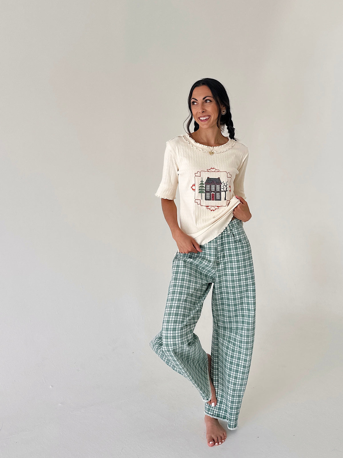 A woman modeling a cream-colored short-sleeve knit top with a graphic print and green-and-white plaid wide-leg pants, standing against a plain light gray studio background.