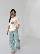 A woman modeling a cream-colored short-sleeve top with a cottage house graphic print and green-and-white plaid wide-leg pants, standing against a plain light gray studio background.