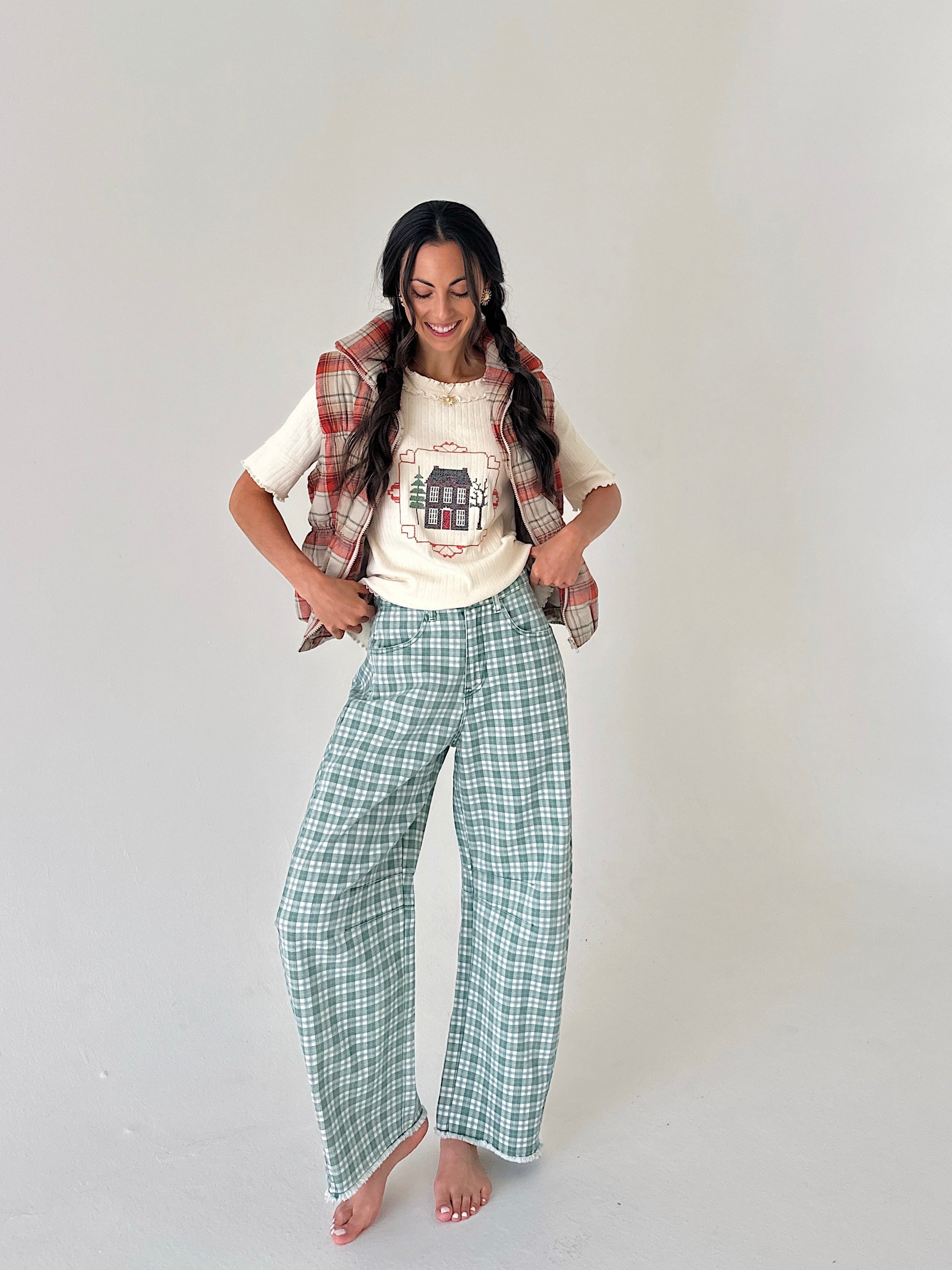 A woman modeling a cozy outfit consisting of wide-leg green and white plaid pants, a cream graphic t-shirt, and a red and green plaid flannel vest draped over her shoulders, set against a plain light gray studio background.