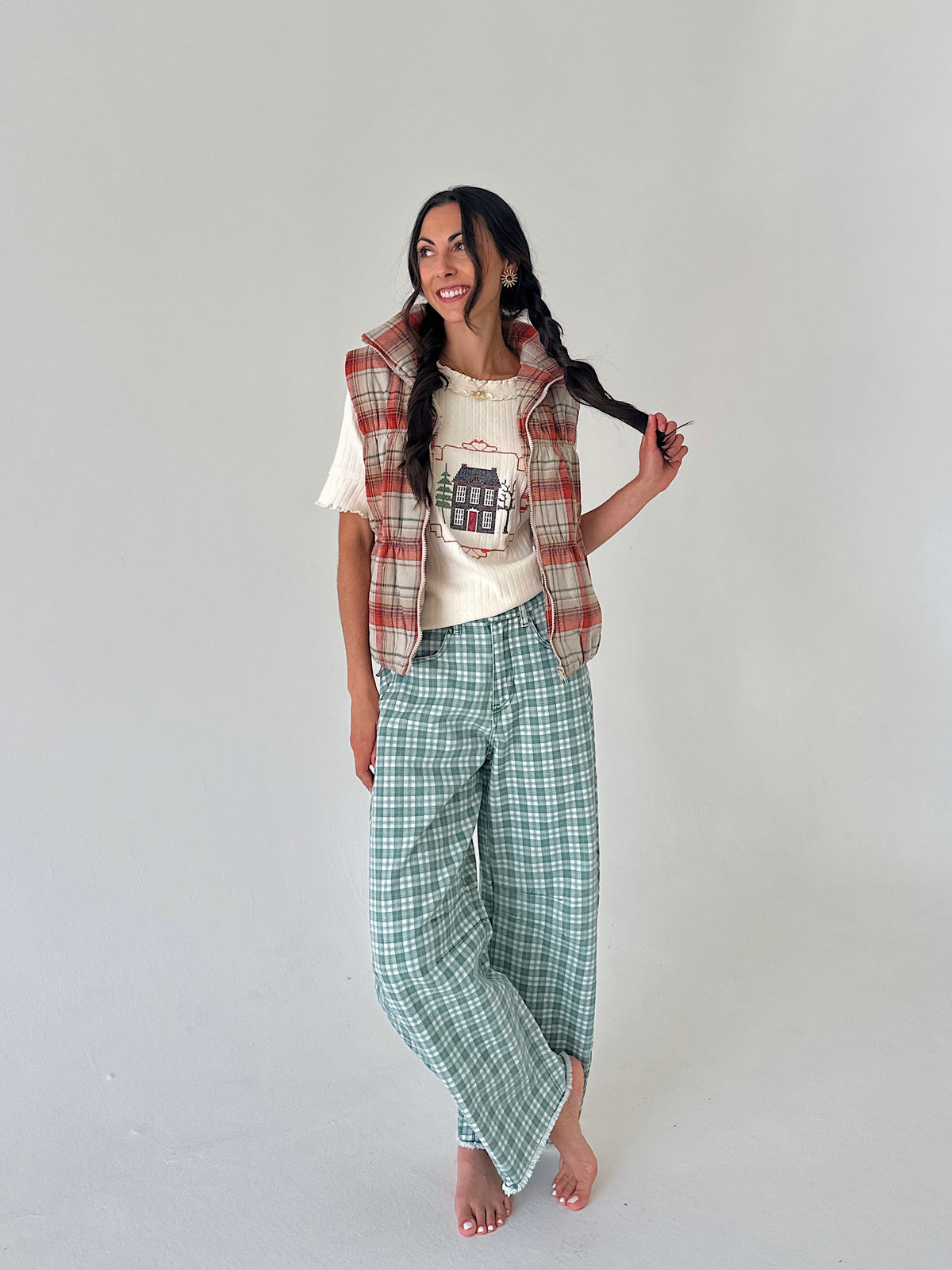 A woman stands barefoot against a plain light gray studio background, wearing a cream graphic T-shirt, a red and beige plaid sleeveless vest, and green-and-white plaid cropped pants.